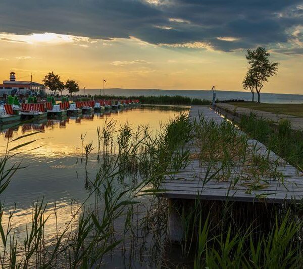 Ein hölzerner, von Gras durchwachsener Steg erstreckt sich bei Sonnenuntergang über das ruhige Wasser, wobei Tretboote und Bäume entlang des Ufers unter einem bewölkten Himmel zu sehen sind.