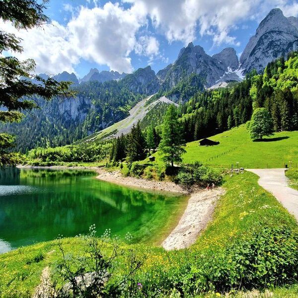 Ein klarer See mit grünem Wasser, gesäumt von Gras und Bäumen, ein Kiesweg, Holzhütten und Berge im Hintergrund unter einem teilweise bewölkten Himmel.