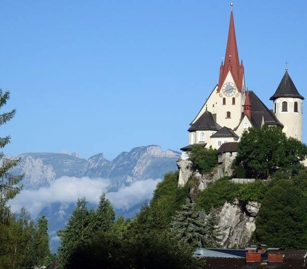 Eine Kirche mit einem hohen roten Kirchturm steht auf einem Hügel, umgeben von Bäumen, mit Bergen und Wolken im Hintergrund unter einem klaren blauen Himmel.