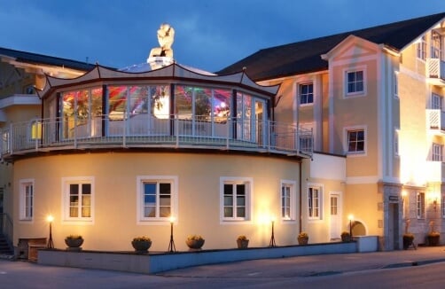 Ein zweistöckiges gelbes Gebäude mit großen Fenstern, einer überdachten Terrasse im Obergeschoss und sanfter Außenbeleuchtung in der Abenddämmerung.