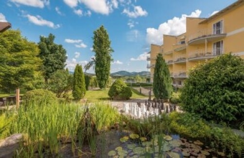 Ein angelegter Garten mit einem kleinen Teich und Seerosen, Bäumen und einem gelben mehrstöckigen Gebäude mit Balkonen unter einem teilweise bewölkten Himmel.