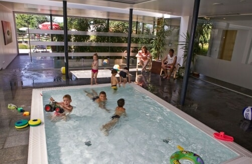 Mehrere Kinder spielen in einem kleinen Hallenbad mit Spielzeug, während Erwachsene in der Nähe auf Bänken sitzen. Helles Licht fällt durch große Fenster ein.