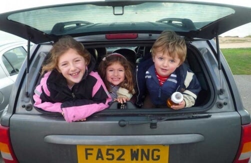 Drei kleine Kinder lächeln, während sie im offenen Kofferraum eines Autos sitzen. Auf dem Nummernschild des Autos steht FA52 WNG.