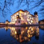 Ein großes, mit leuchtenden Sternen geschmücktes Gebäude spiegelt sich in einem ruhigen Gewässer in der Abenddämmerung, mit kahlen Baumzweigen im Vordergrund.