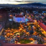 Luftaufnahme eines Stadtplatzes in der Abenddämmerung, beleuchtet von festlichen Lichtern und versammelten Menschenmengen, umgeben von Gebäuden und Bergen im Hintergrund.