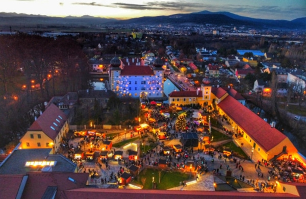 Luftaufnahme eines Stadtplatzes in der Abenddämmerung, beleuchtet von festlichen Lichtern und versammelten Menschenmengen, umgeben von Gebäuden und Bergen im Hintergrund.
