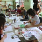 Eine Gruppe von Menschen sitzt in einem Kunstatelier um einen Tisch herum und zeichnet und malt auf Papier, wobei verschiedene Kunstmaterialien auf dem Tisch verstreut sind.