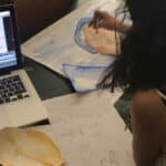 Eine Person sitzt an einem Tisch und skizziert und malt, während sie ein Animationsvideo auf einem Laptop betrachtet, auf dem Storyboards und Kunstwerke ausgebreitet sind.