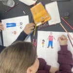 Zwei Kinder sitzen an einem Tisch und zeichnen Bilder von Menschen auf weißem Papier, umgeben von Buntstiften und Malutensilien.