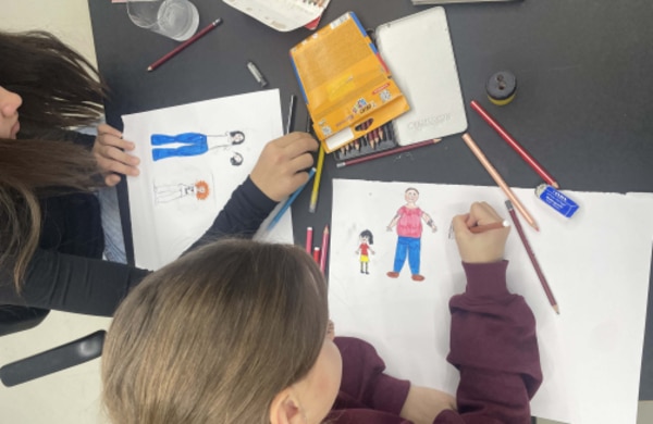 Zwei Kinder sitzen an einem Tisch und zeichnen Bilder von Menschen auf weißem Papier, umgeben von Buntstiften und Malutensilien.