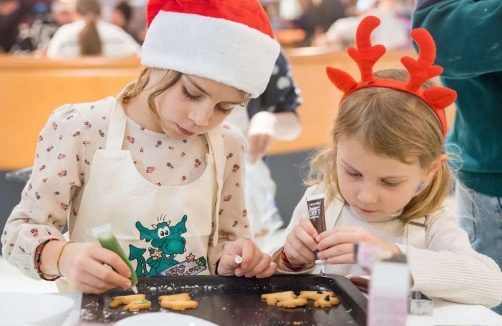 Zwei junge Mädchen verzieren Plätzchen auf einem Backblech. Die eine trägt eine Weihnachtsmannmütze und die andere ein Rentiergeweih.