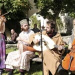 Vier Musiker spielen im Freien; einer spielt Geige, einer Trommel, einer Oud und einer Cello. Sie sitzen im Gras vor Bäumen und Menschen im Hintergrund.