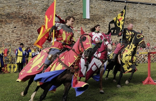 Drei Personen in mittelalterlichen Ritterkostümen reiten auf Pferden, die mit passenden heraldischen Bannern geschmückt sind, während eines Ritterturniers in der Nähe einer Steinmauer.