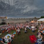 Eine große Menschenmenge sitzt auf Stühlen und Decken und sieht sich in der Abenddämmerung einen Freiluftfilm vor einer historischen Steinmauer an, an deren Seite ein Zelt aufgebaut ist.