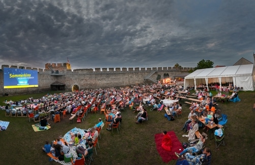 Eine große Menschenmenge sitzt auf Stühlen und Decken und sieht sich in der Abenddämmerung einen Freiluftfilm vor einer historischen Steinmauer an, an deren Seite ein Zelt aufgebaut ist.