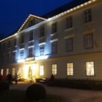 Ein großes, dreistöckiges, mit Lichterketten geschmücktes Gebäude in der Abenddämmerung. Mehrere Menschen sind in der Nähe eines Eingangs mit der Aufschrift "ADVENT" versammelt.