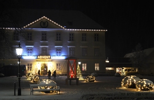 Großes Gebäude, das nachts mit Weihnachtslichtern geschmückt ist, mit beleuchteten Büschen und einer roten Struktur davor, Menschen betreten einen Eingang mit der Aufschrift "Advent". Schnee bedeckt den Boden.