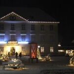 Ein großes Gebäude, das mit Lichtern und einem "ADVENT"-Schild geschmückt ist; beleuchtete Skulpturen und Bäume sind nachts in einem verschneiten Innenhof zu sehen.