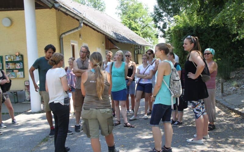 Eine Gruppe von Erwachsenen steht im Freien in einem Halbkreis in der Nähe eines Gebäudes und hört einer Person zu, die spricht. Einige Personen tragen Sonnenbrillen und Sommerkleidung. Bäume und eine Anzeigetafel sind zu sehen.